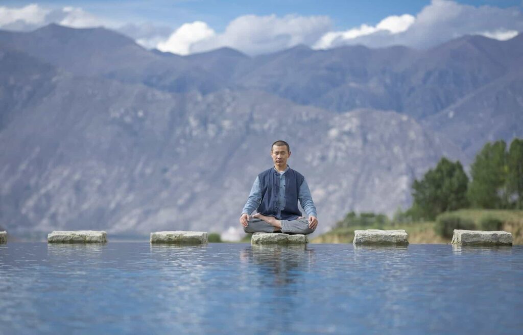 Man meditating on stepping stones in middle of river - Vipassana Meditation